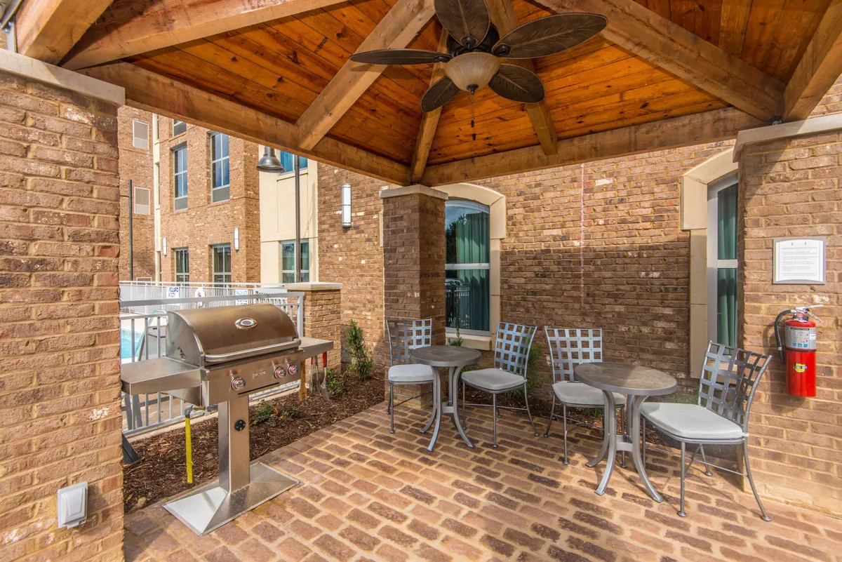 An inviting exterior scene featuring a covered seating area with comfortable furniture, a grill setup, and a wooden ceiling overhead. The area is ideal for outdoor gatherings, with sunlight casting a warm glow and plenty of space for relaxation or socializing.