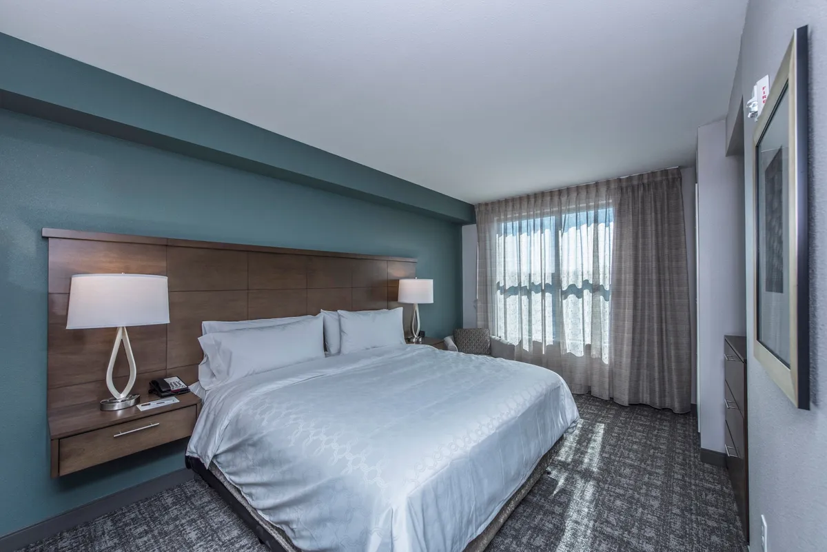 A tranquil bedroom scene with a king-sized bed dressed in crisp linens, flanked by a nightstand and lamp, and large windows that flood the space with soothing natural light. The overall mood is peaceful and restful, ideal for unwinding or taking a nap.