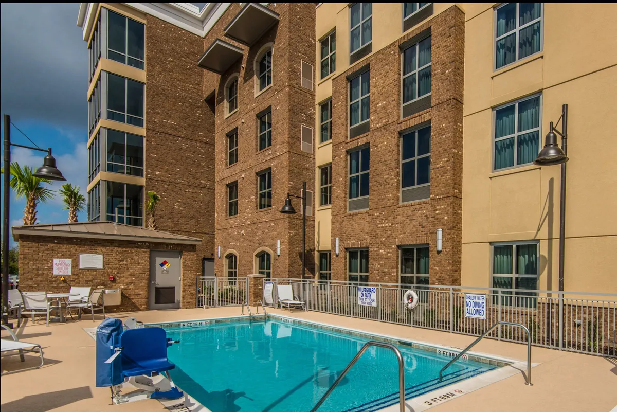 A sparkling outdoor swimming pool is surrounded by palm trees, lounge chairs, and the soft evening light, with the building’s facade in the background. The inviting setting hints at leisurely daytime swims and relaxing moments under the sun.