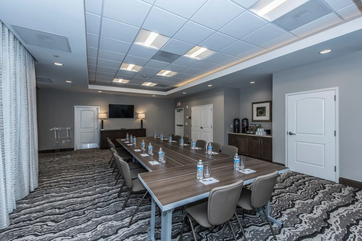 A sleek, professional conference room with a long table, water bottles, a TV screen for presentations, and decorative wall art. The setting is designed for focused meetings, workshops, or group projects.