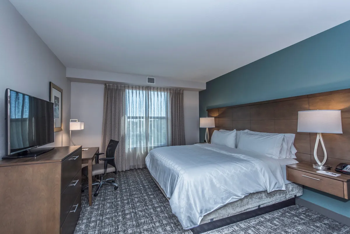 A serene bedroom with a luxurious king-sized bed, a TV positioned for comfortable viewing, a window with flowing curtains that lets in natural light, a desk and chair for work or reading, and a nightstand with a soft-lit lamp. The decor is tranquil and inviting, designed for both relaxation and productivity.