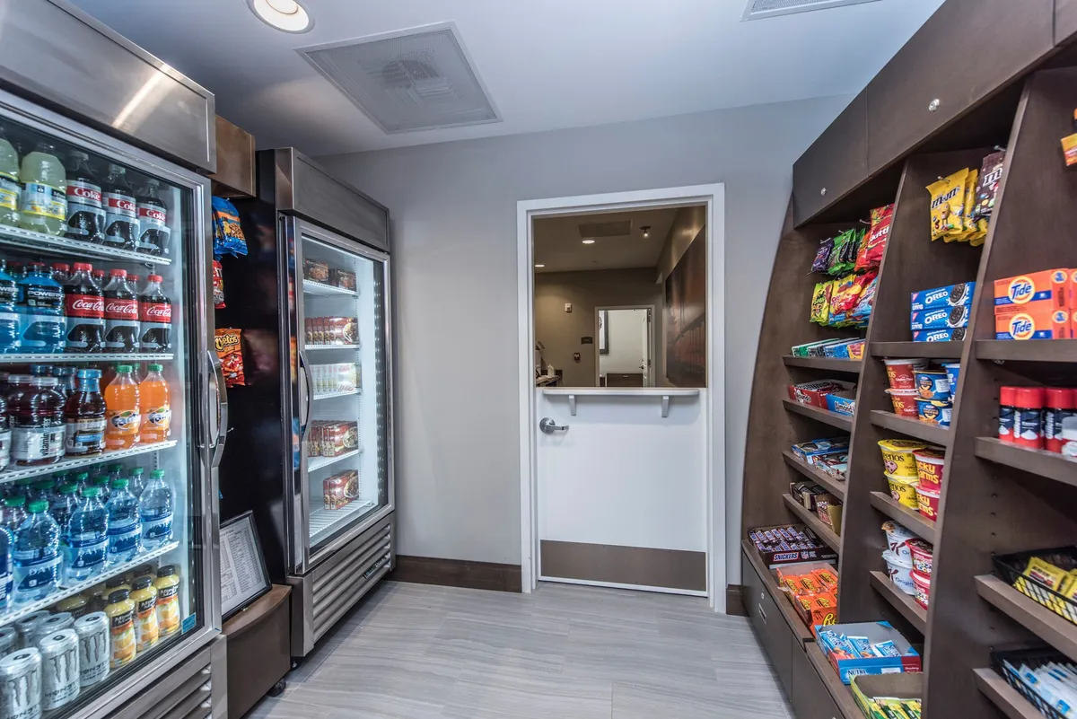 A casual snack area featuring shelves stocked with a variety of snacks and a refrigerated case filled with cold drinks. An exit door is visible, suggesting quick access for guests on the go, while the space invites light social interaction and convenient refreshment.