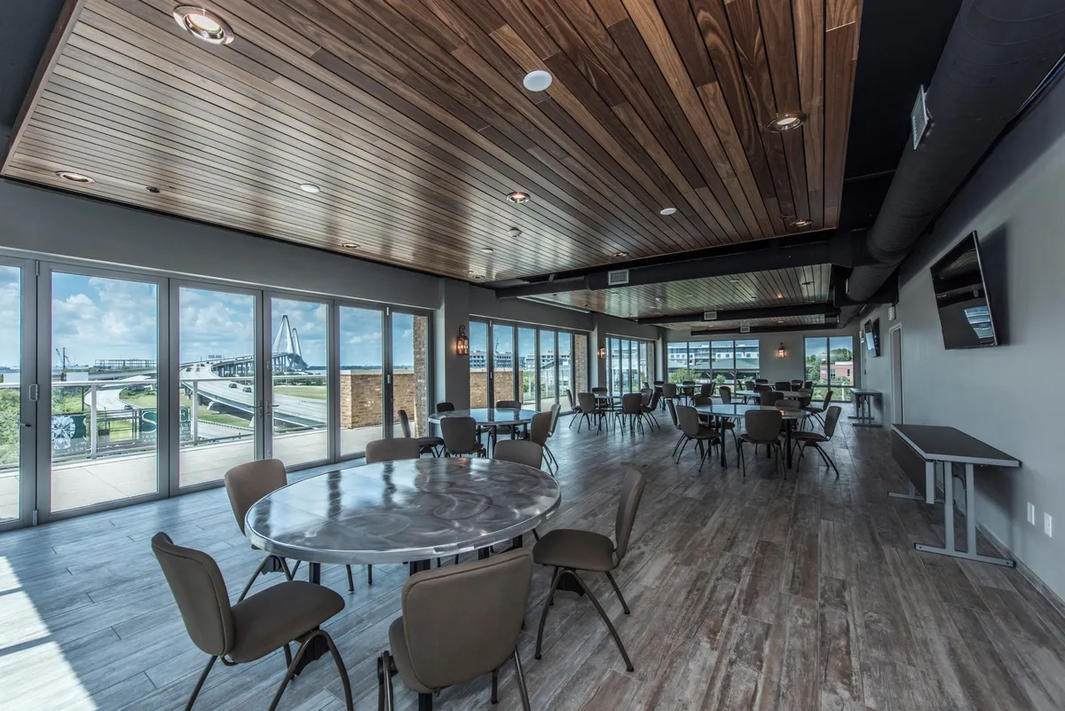 A bright, airy interior common area with large windows offering scenic views, round tables for dining or meetings, and a stylish wooden ceiling. The atmosphere is welcoming and designed for both casual socializing and informal work sessions.