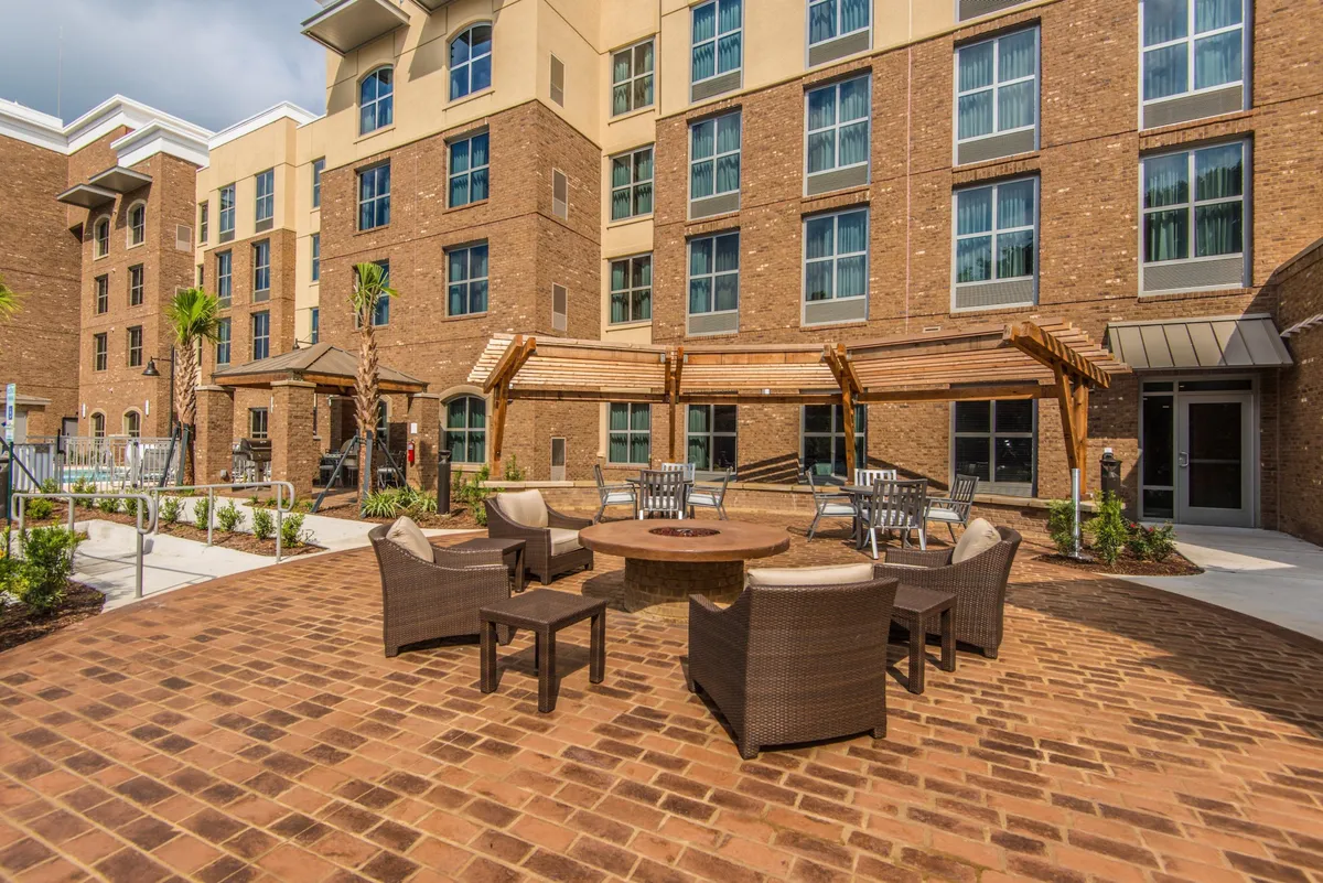 A cozy outdoor fire pit area with comfortable seating, palm trees, and the building’s architecture visible behind. The scene suggests both daytime and evening social gatherings in a welcoming environment.