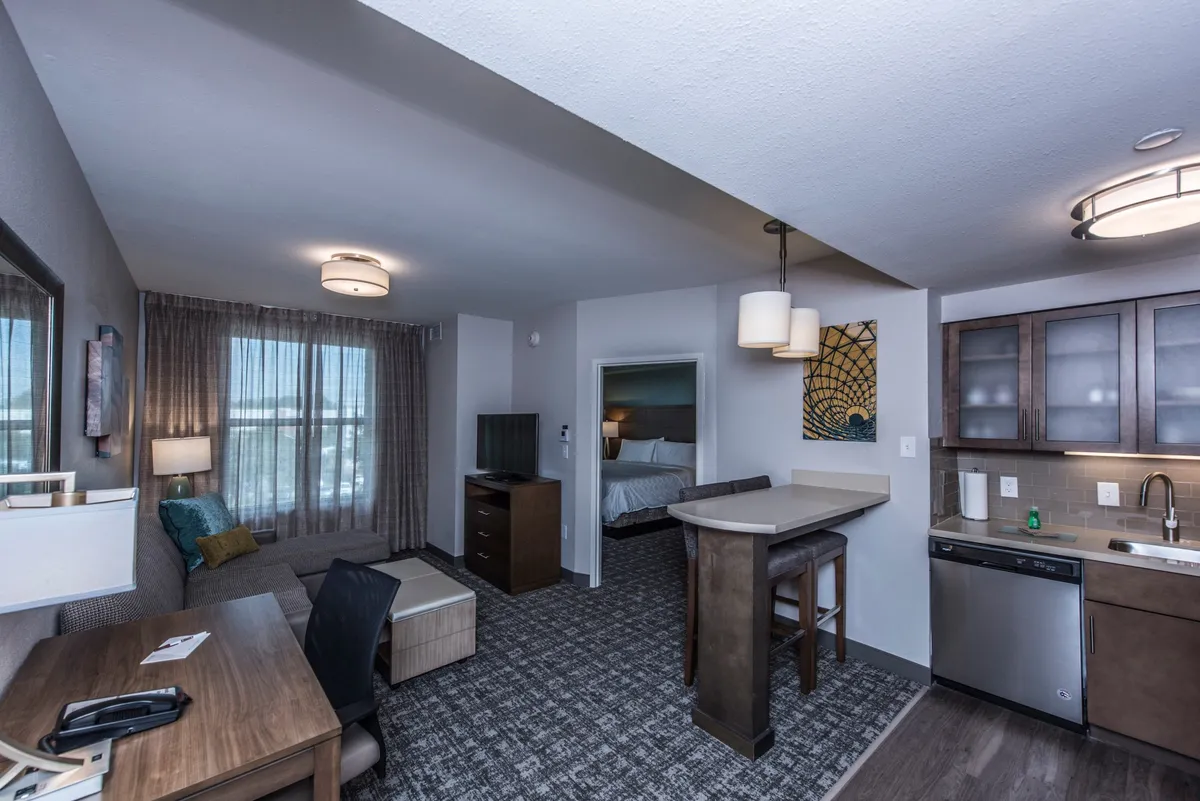 The image showcases a cozy condo interior featuring a plush sofa adorned with colorful cushions, a wooden desk ideal for work or planning adventures, a modern bar counter with stools perfect for casual dining, and a fully equipped kitchenette. Natural light pours in from the window, adding warmth to the inviting setting.