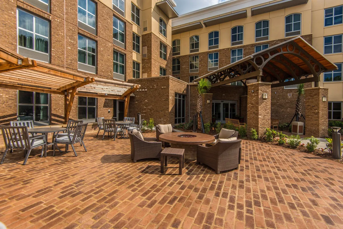 An inviting fire pit surrounded by a comfortable seating area on a brick patio, with palm trees and the hotel’s facade in the background. The space is clearly designed for social gatherings and relaxation, especially during the evening as the sun sets.