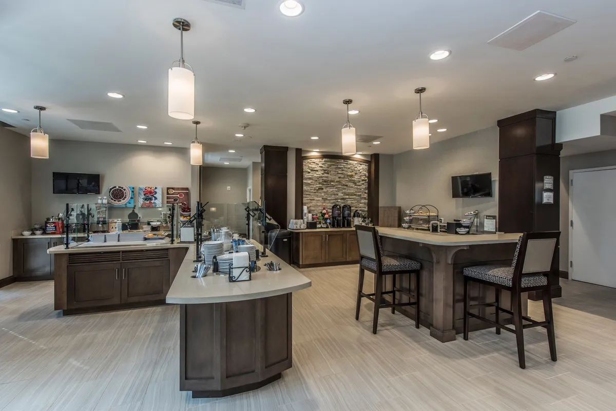 A bright, inviting breakfast area featuring a central countertop with a coffee station and ample seating. The décor includes a decorative stone accent wall and warm lighting, creating a welcoming place for guests to gather for complimentary breakfast or relax with a coffee.