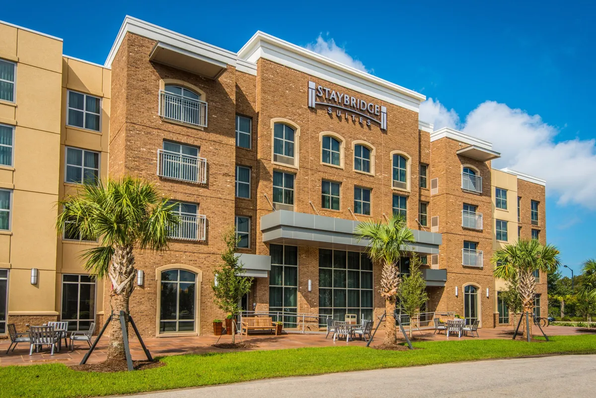 This exterior image features the Staybridge Suites sign set against large windows, a patio with outdoor seating, and lush palm trees, all bathed in inviting sunlight. The welcoming atmosphere is evident as people relax outdoors, suggesting both approachability and comfort.